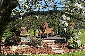 Im Vordergrund ein blühender Baum, dahinter eine Ruheinsel in einem von einer Hecke begrenzten Garten mit zwei Liegen und zwei Skulpturen mit Gesichtern